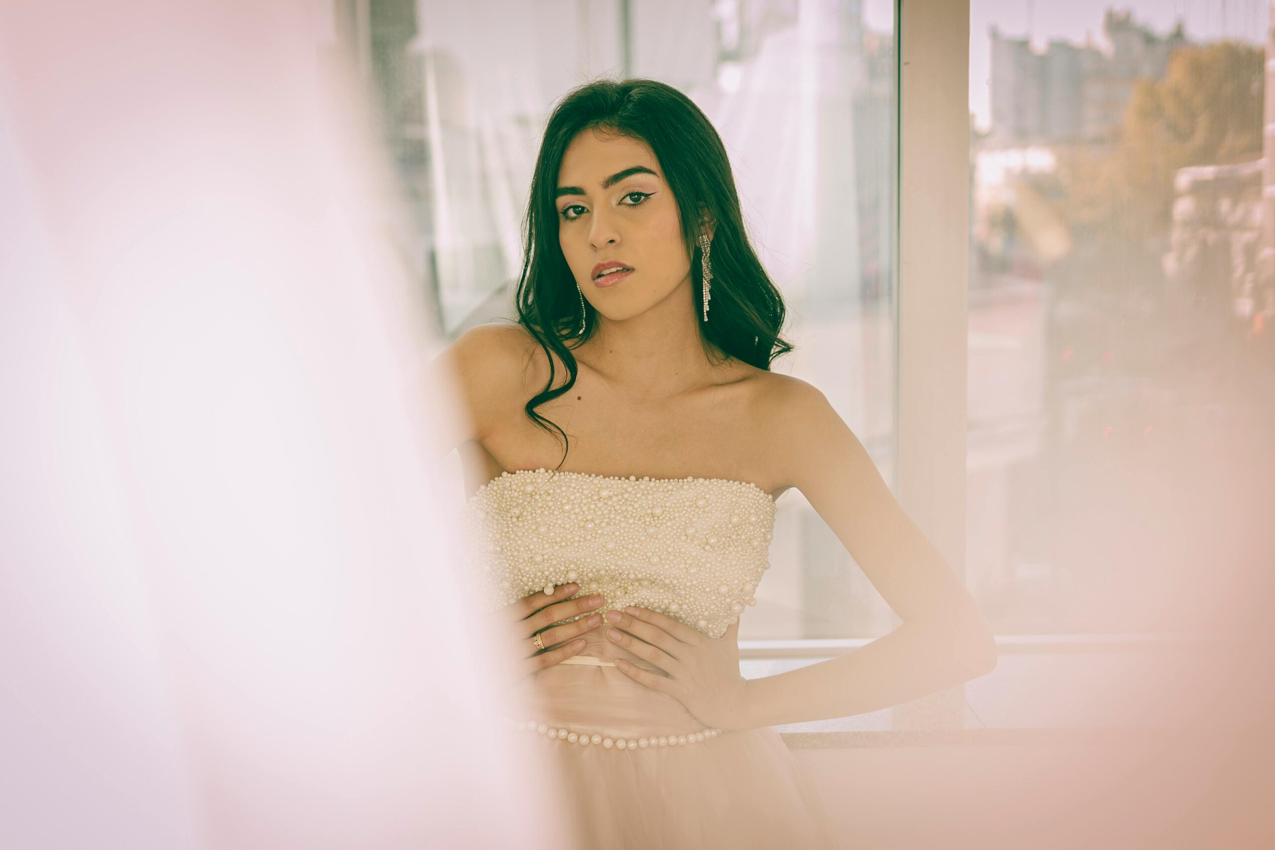 A young woman poses gracefully in a pearl-embellished dress with a soft, dreamy look by a window in Buenos Aires.